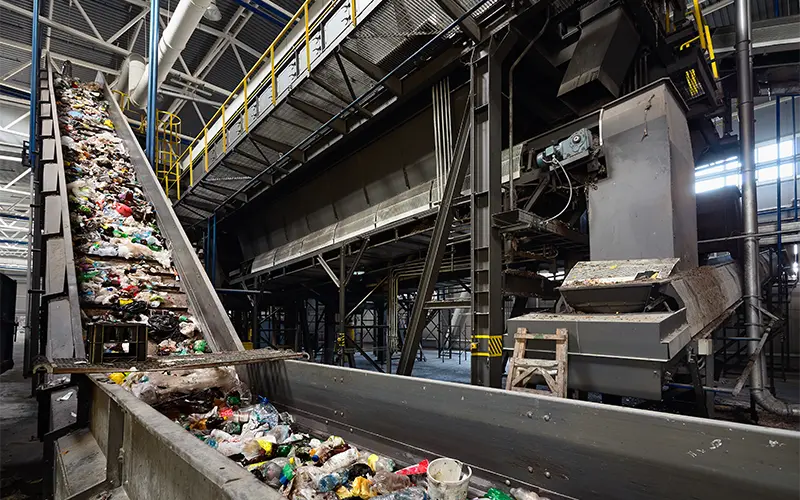 Conveyor belt at recycling plant transports garbage inside drum filter or rotating cylindrical sieve with trommel or screen for sorting pieces of garbage into various sizes fractions.